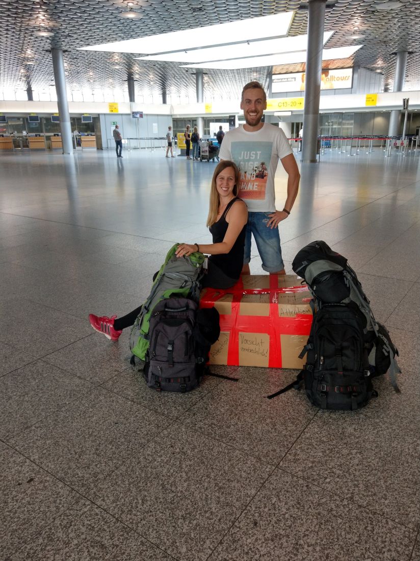 Malina und ihr Mann am Flughafen mit Rucksäcken und Umzugskartons, kurz vor dem Aufbruch in ein neues Leben