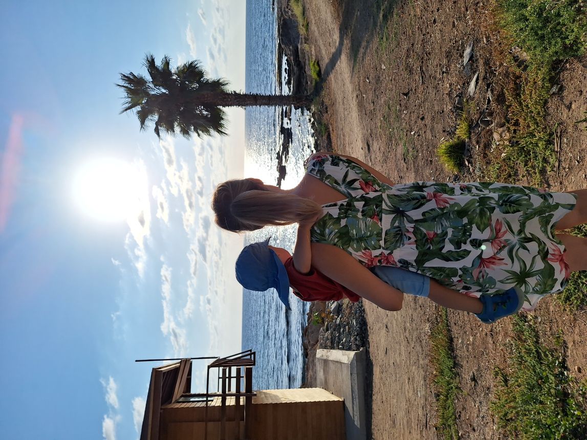 Malina hält ihr Kind im Arm an einem sonnigen Küstenort mit Palme und Meer